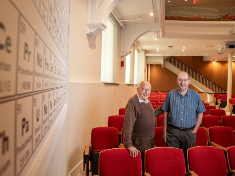 professor, alum stand in row of red chairs