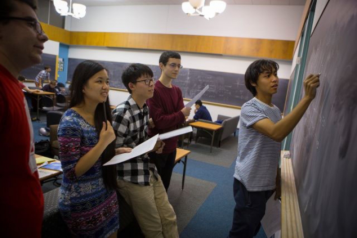 Four students standing behind a teaching assistant who is writing on a chalk board