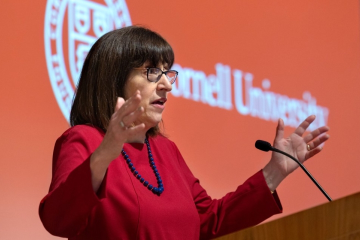 The Cornell President, a brunette woman, stands in a red blazer at a lectern