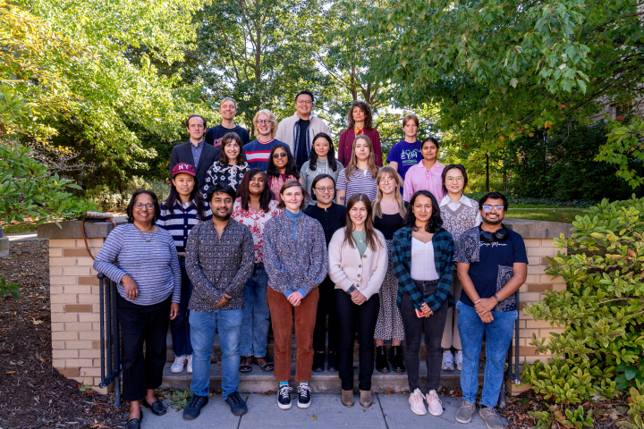 A group of twenty graduate students gather for a group photo under green trees in summer.